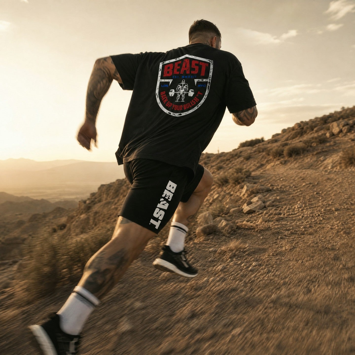 A man running on a desert mountain trail wearing a black t-shirt and black shorts with 'BEAST' branding, photographed from behind in warm golden hour light.